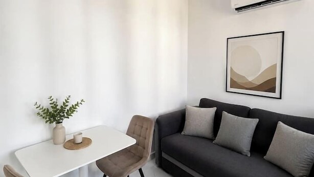 A modern living room with a white table, two chairs, a dark sofa, a wall-mounted air conditioner, and a framed artwork. - Elegant Urban Retreat | Near Old Town (Bucharest)