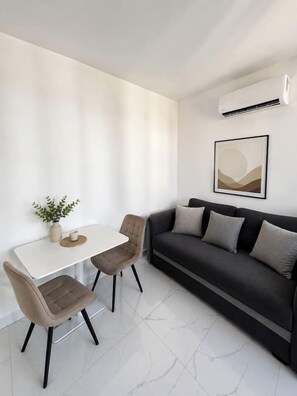 Een moderne woonkamer met een witte tafel, twee stoelen, een donkere bank, een wandairconditioner en een lijstwerk met kunst.