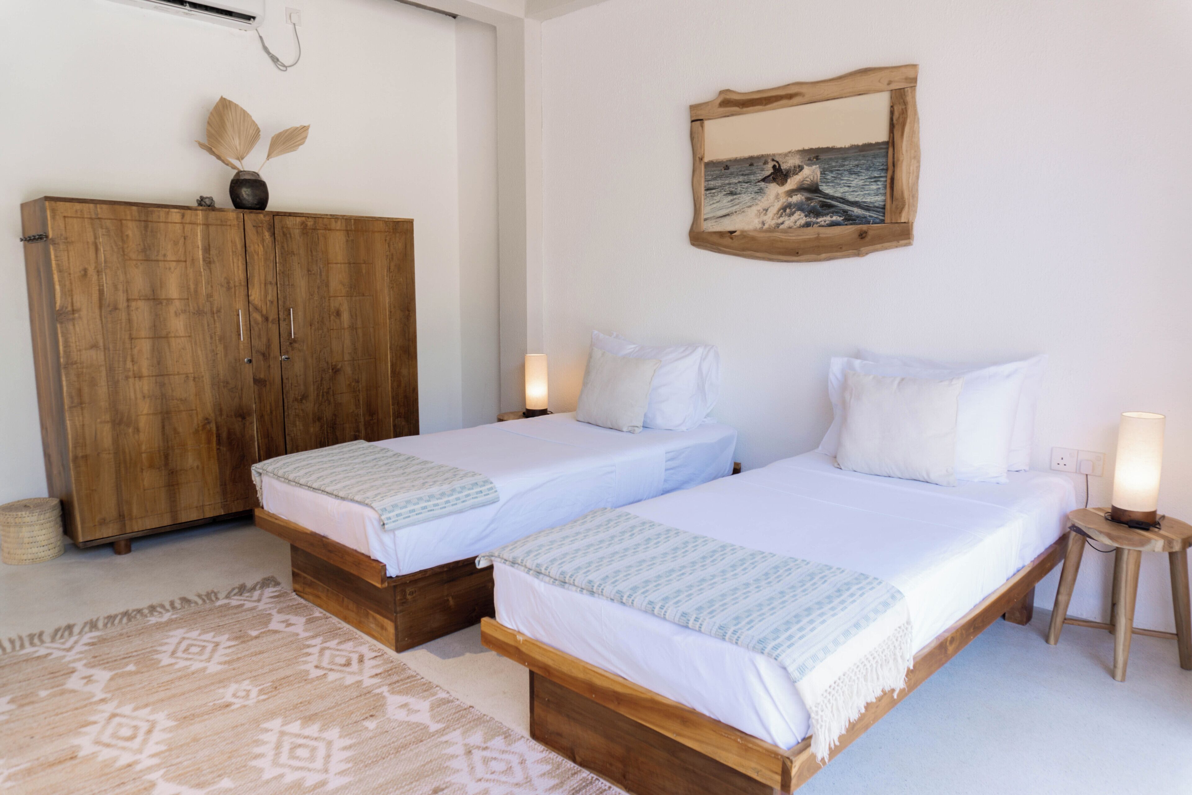 Een kamer met twee enkele bedden, een houten garderobe, een lijst met een afbeelding aan de muur, en een tafeltje met een lamp.
