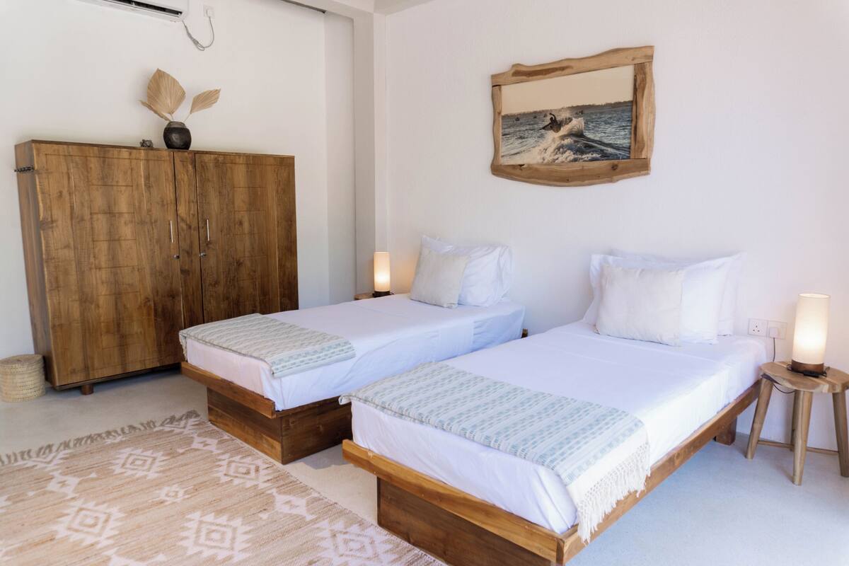 A room with two single beds, a wooden wardrobe, a framed picture on the wall, and a small table with a lamp.