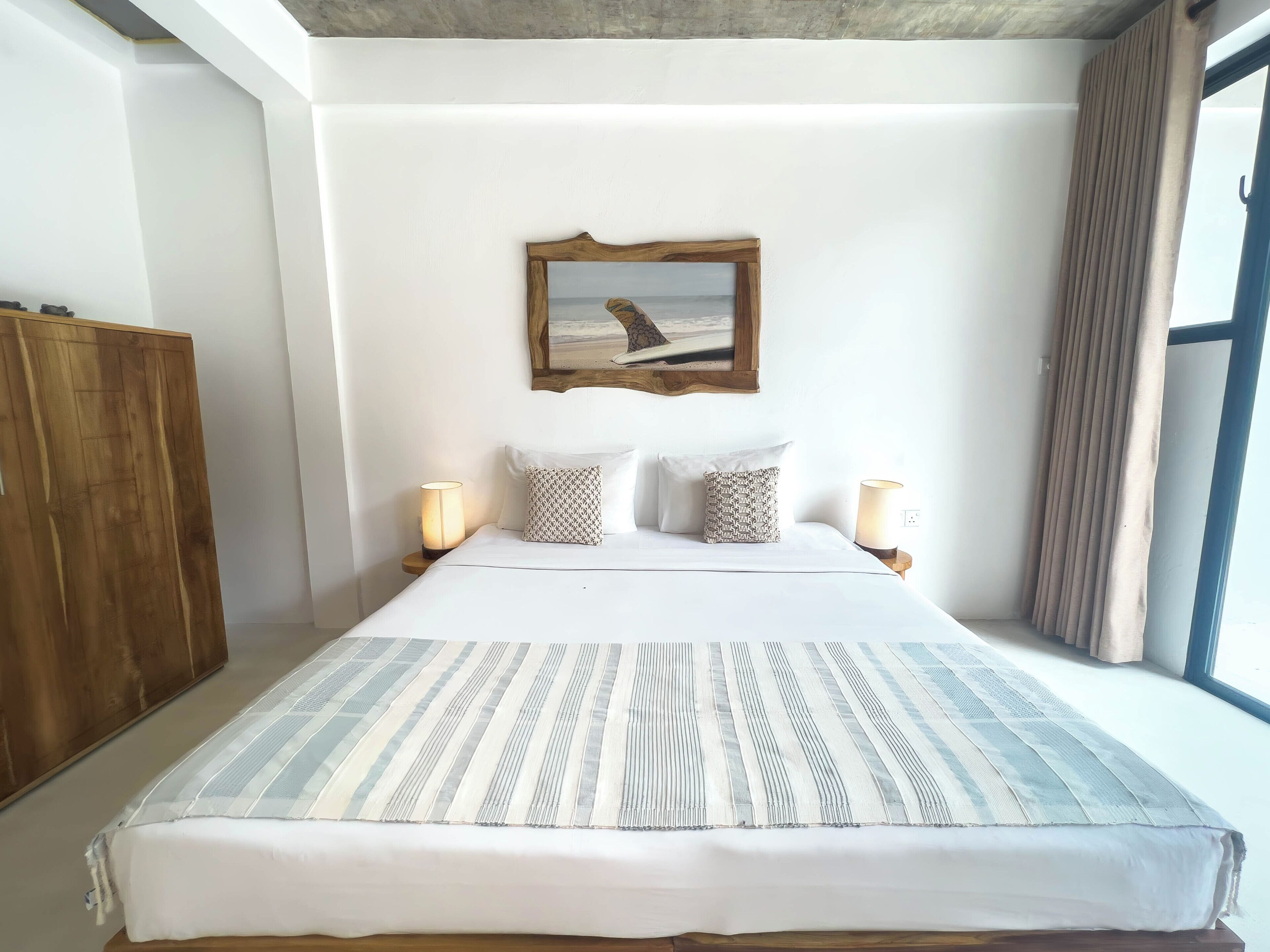 A hotel room with a large bed, two bedside lamps, a framed picture on the wall, and a window with curtains.