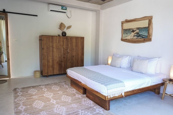 A bedroom with a wooden bed, white bedding, a patterned rug, a wooden wardrobe, and a framed picture on the wall.
