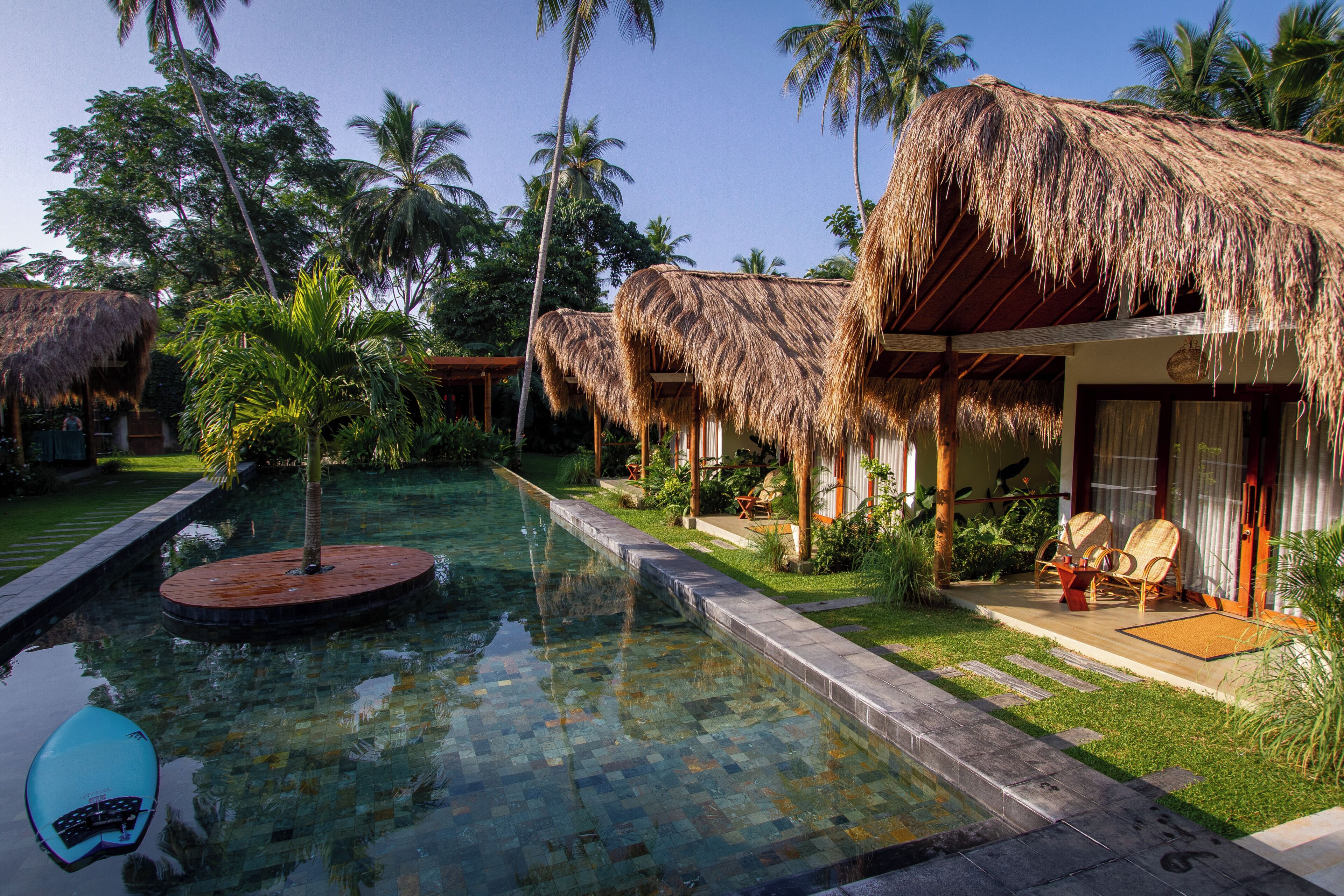 Un área de piscina con construcciones de techo de paja, palmeras y una piscina cristalina.