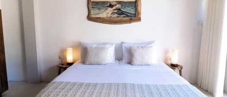 Un lit bien fait, avec du linge de lit blanc et une couverture à motifs, flanqué de deux lampes de chevet posées sur des tables de nuit en bois. Un tableau encadré représentant un paysage marin est accroché au-dessus du lit.
