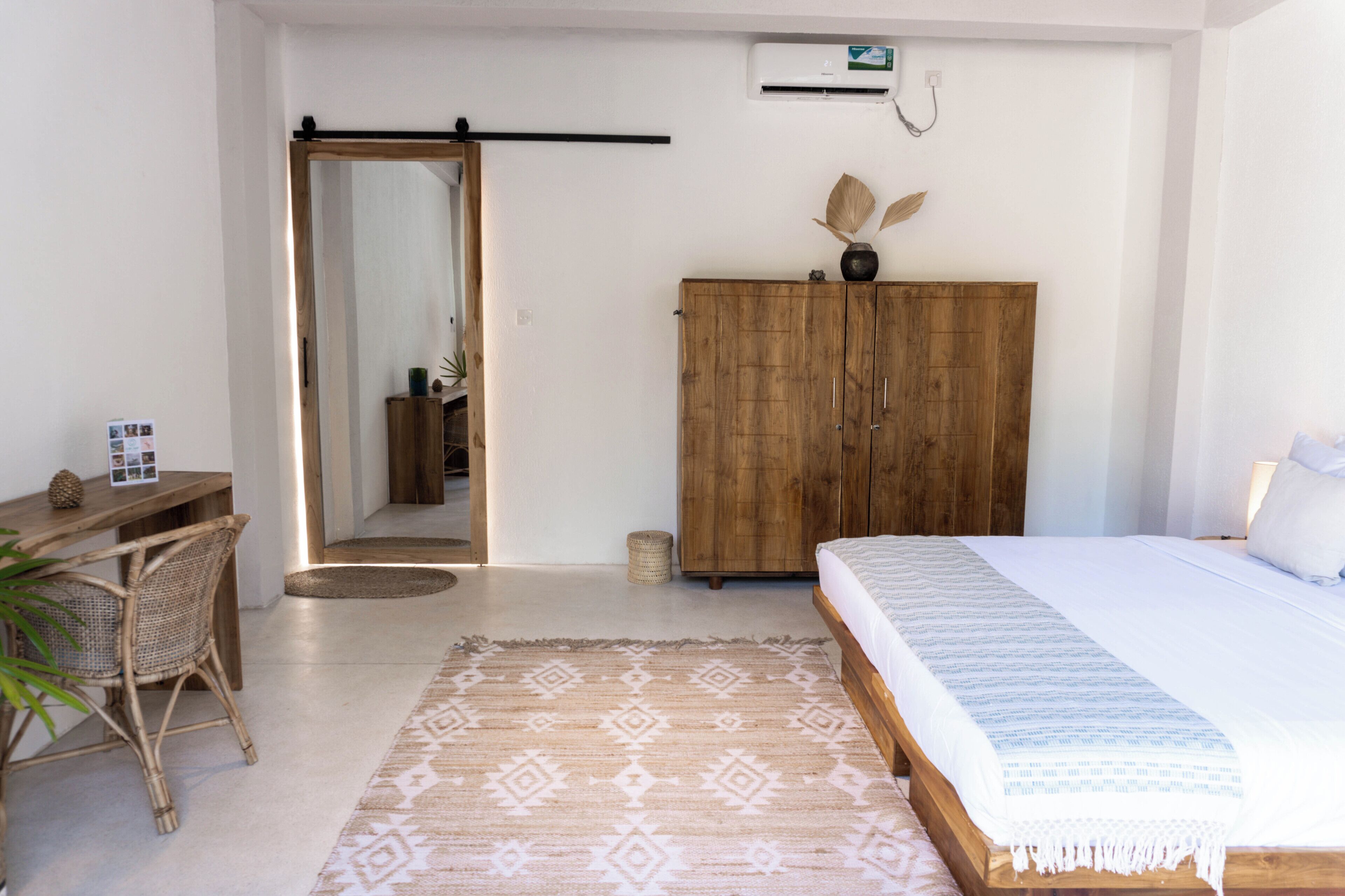 A bedroom with a bed, a wooden wardrobe, a desk with a chair, and a mirror.