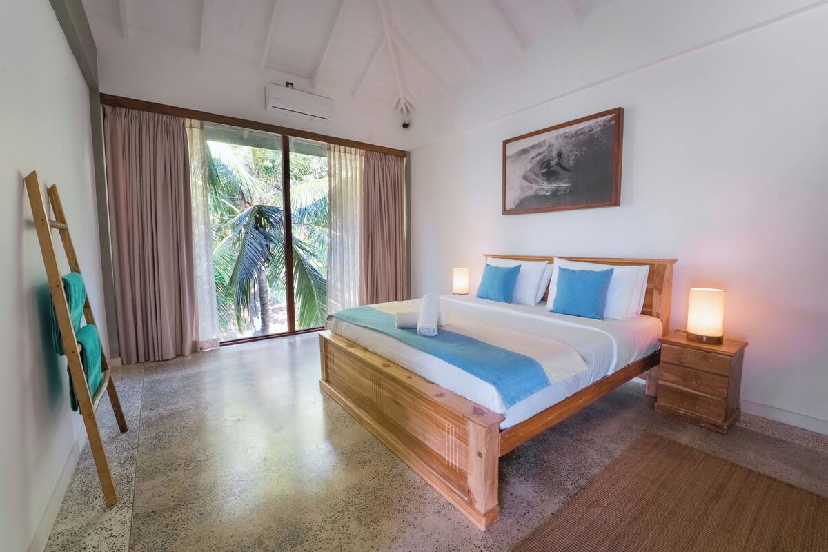 Un dormitorio con una cama de madera, una mesita de noche con una lámpara, un cuadro en la pared y vistas a palmeras a través de la ventana.