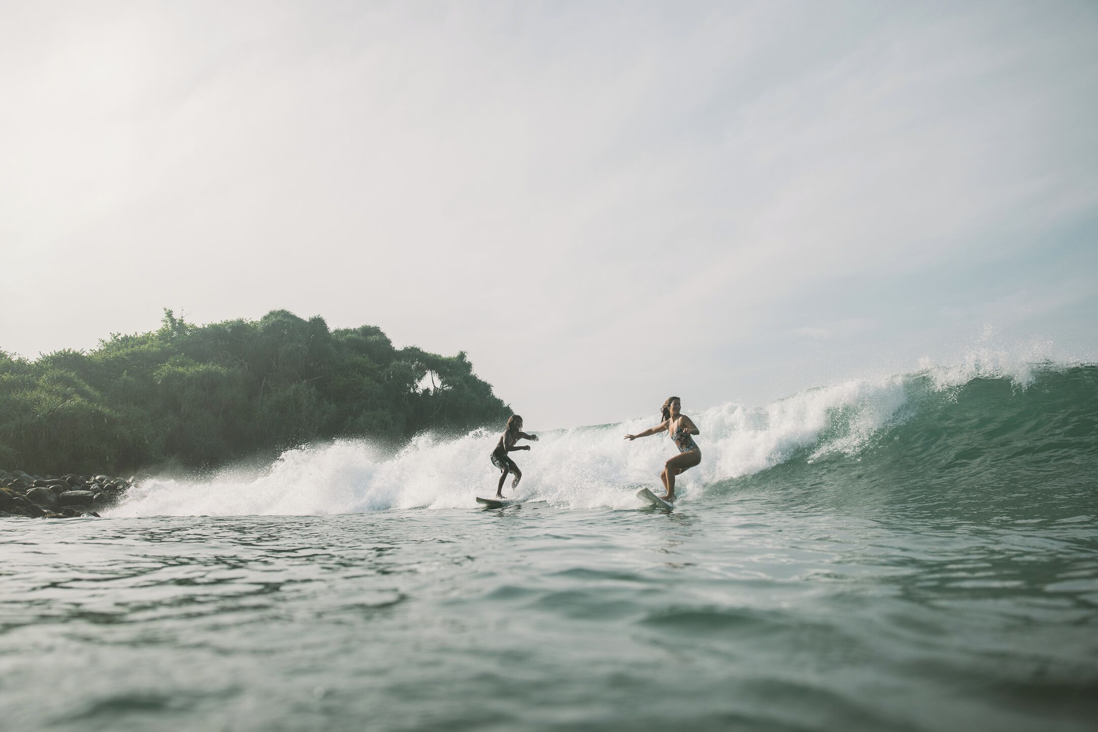 Two individuals surfing on ocean waves with a forested area in the background.