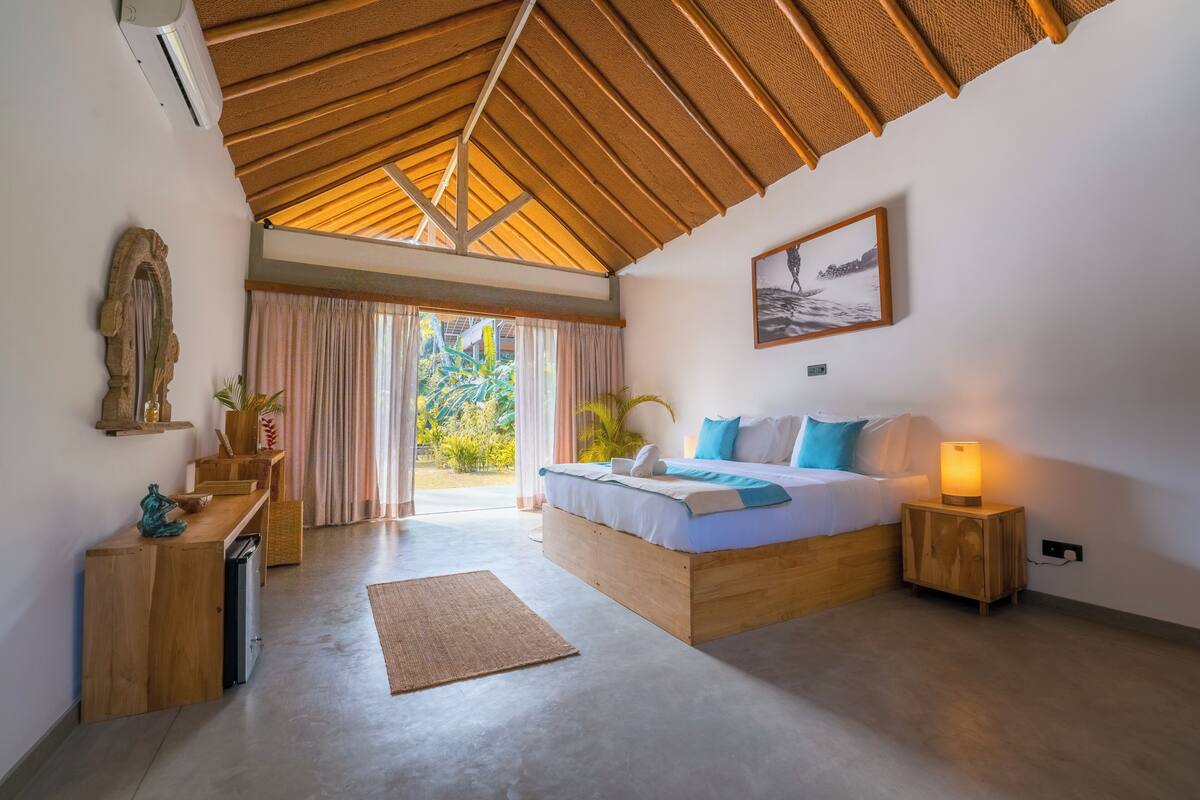 Un dormitorio con techo de madera, una cama con almohadas, una lámpara de noche, un pequeño mueble de madera y un gran ventanal con cortinas.