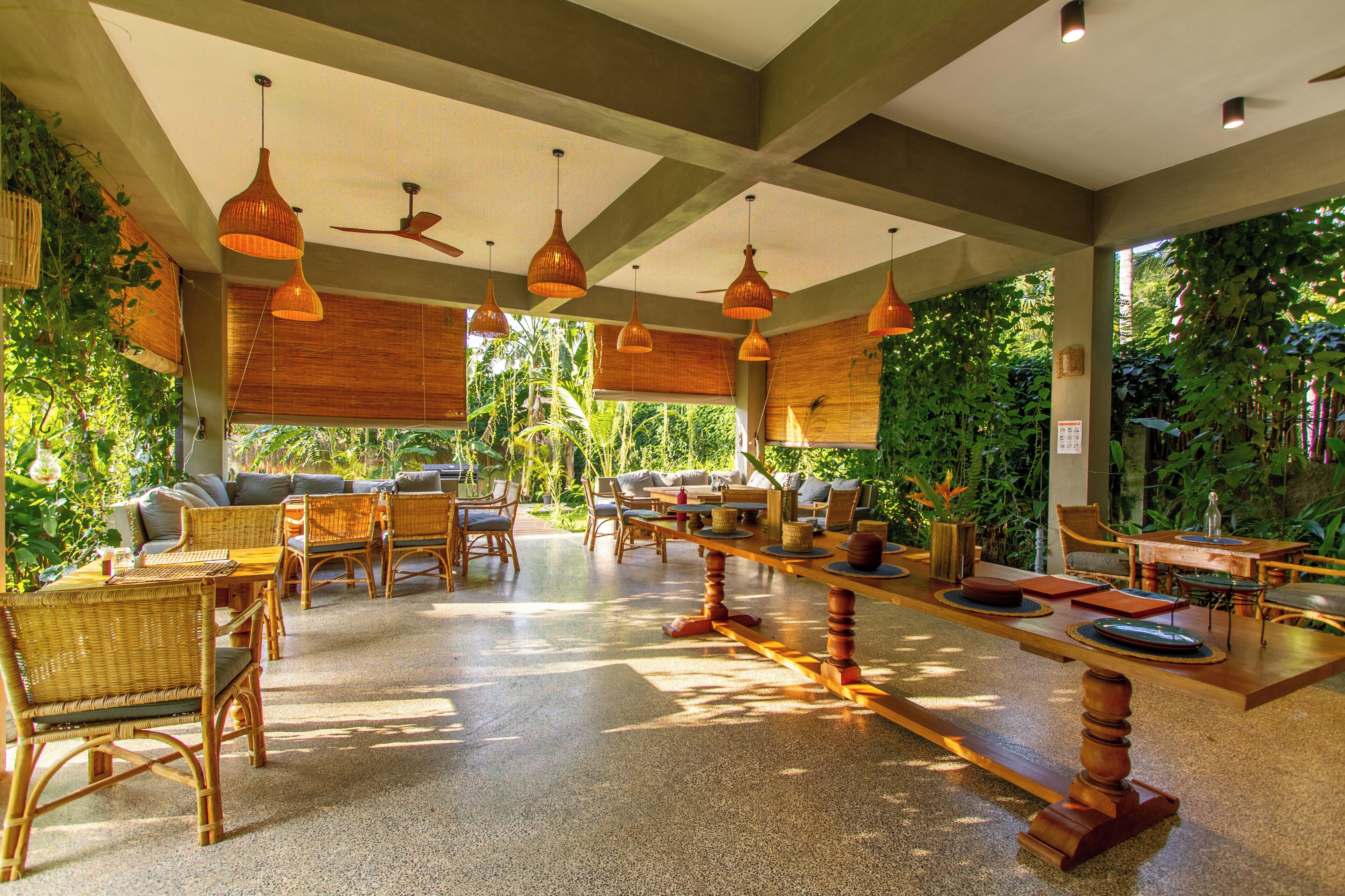 A spacious indoor area with a long wooden table, wicker chairs, and hanging pendant lights.
