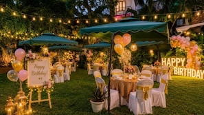 Decoración para una fiesta de cumpleaños al aire libre, con mesas, sillas y luces colgantes.