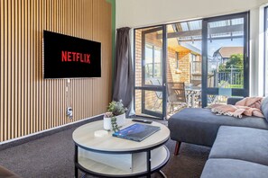 Una sala de estar moderna con televisor de pantalla plana montado en una pared de madera, un sofá, una mesa de centro y vistas a un balcón con mobiliario exterior.