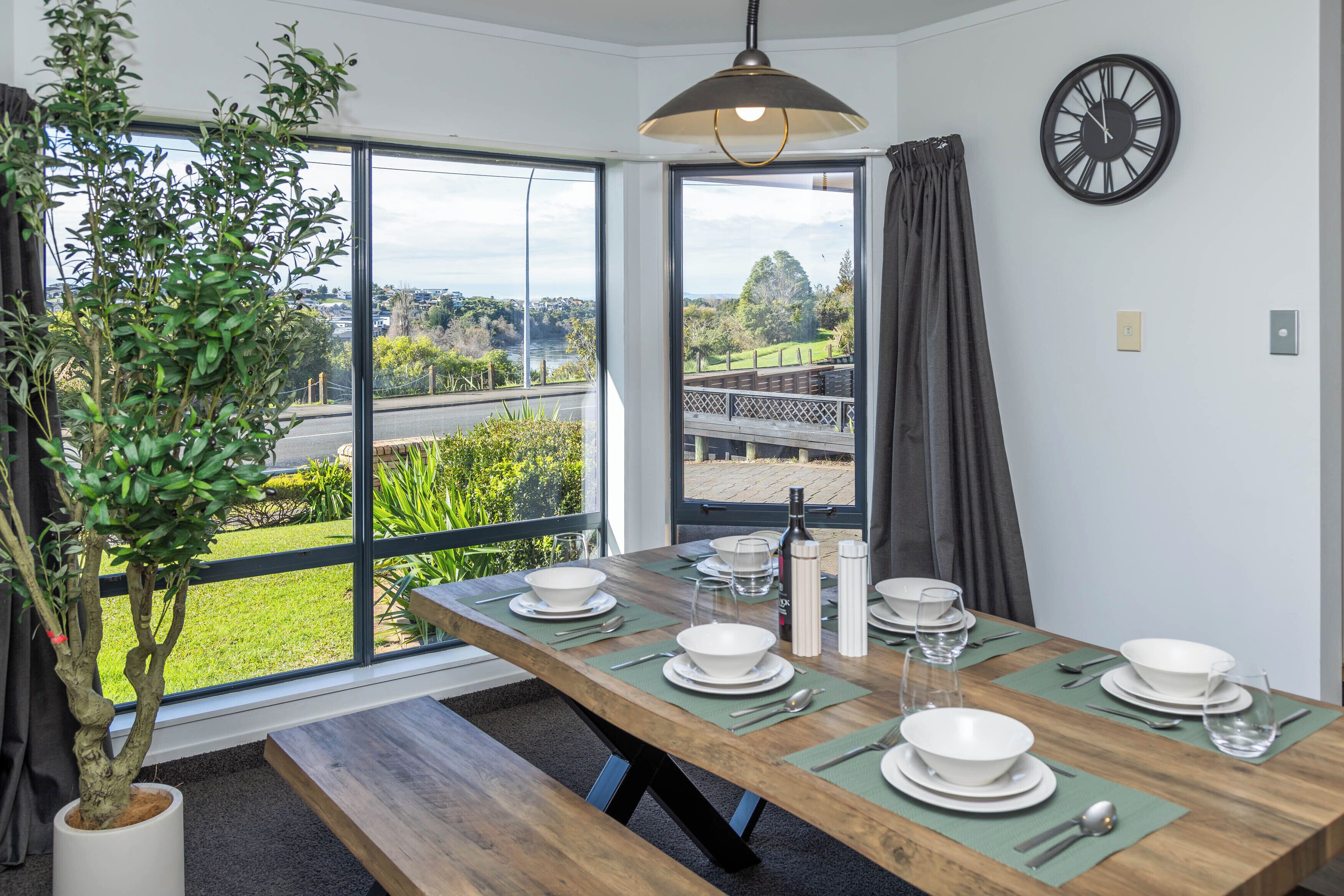 A dining area with a table set for four, a potted plant, a clock, and a view of a river and greenery outside.