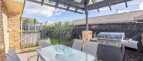 An outdoor patio with a glass table, metal chairs, a barbecue grill, and a brick wall.