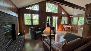 Un intérieur de chalet chaleureux avec une cheminée, un coin salon et de grandes fenêtres laissant entrer la lumière naturelle.