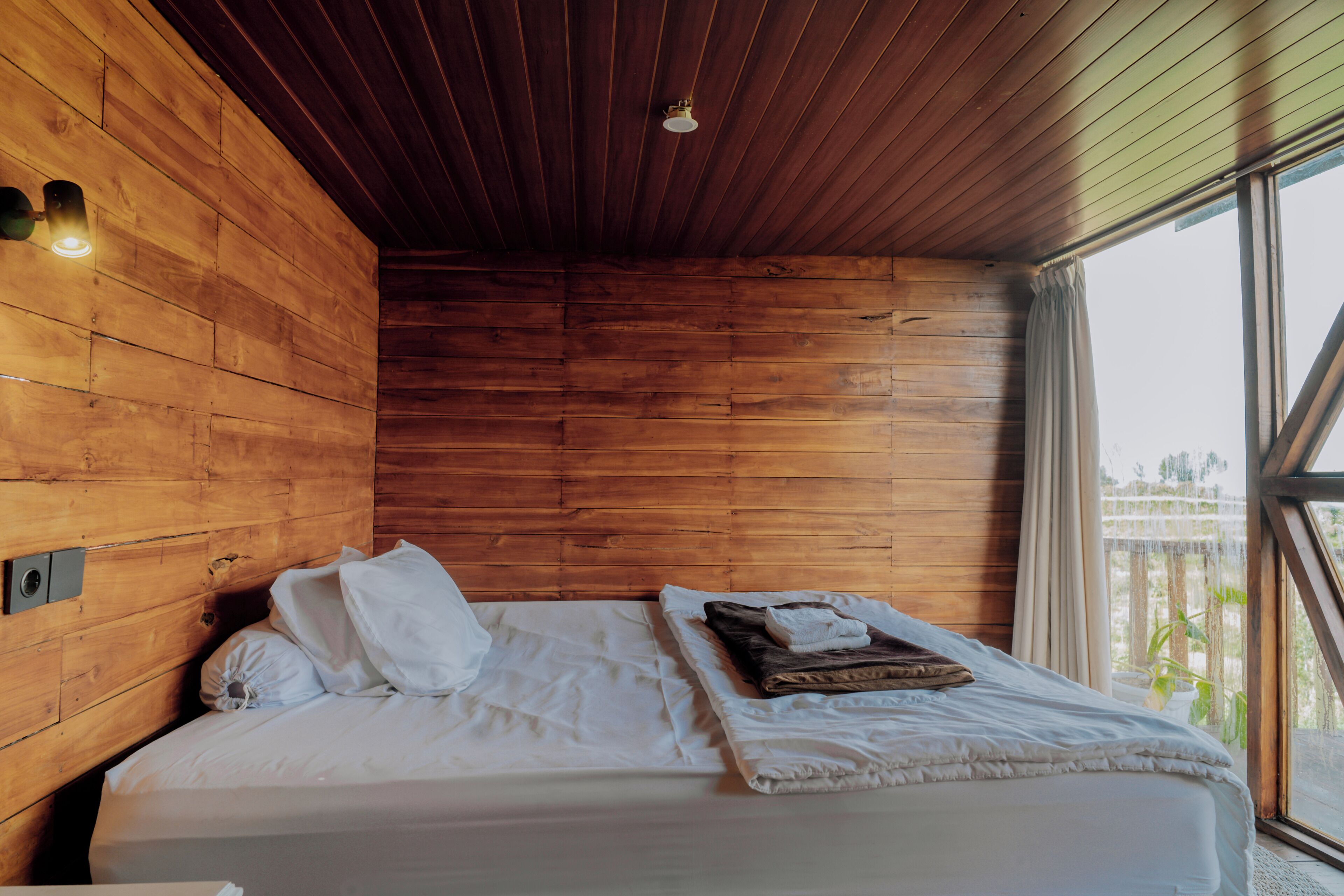 Une pièce aux murs en bois, un lit avec une literie blanche et une fenêtre avec des rideaux.