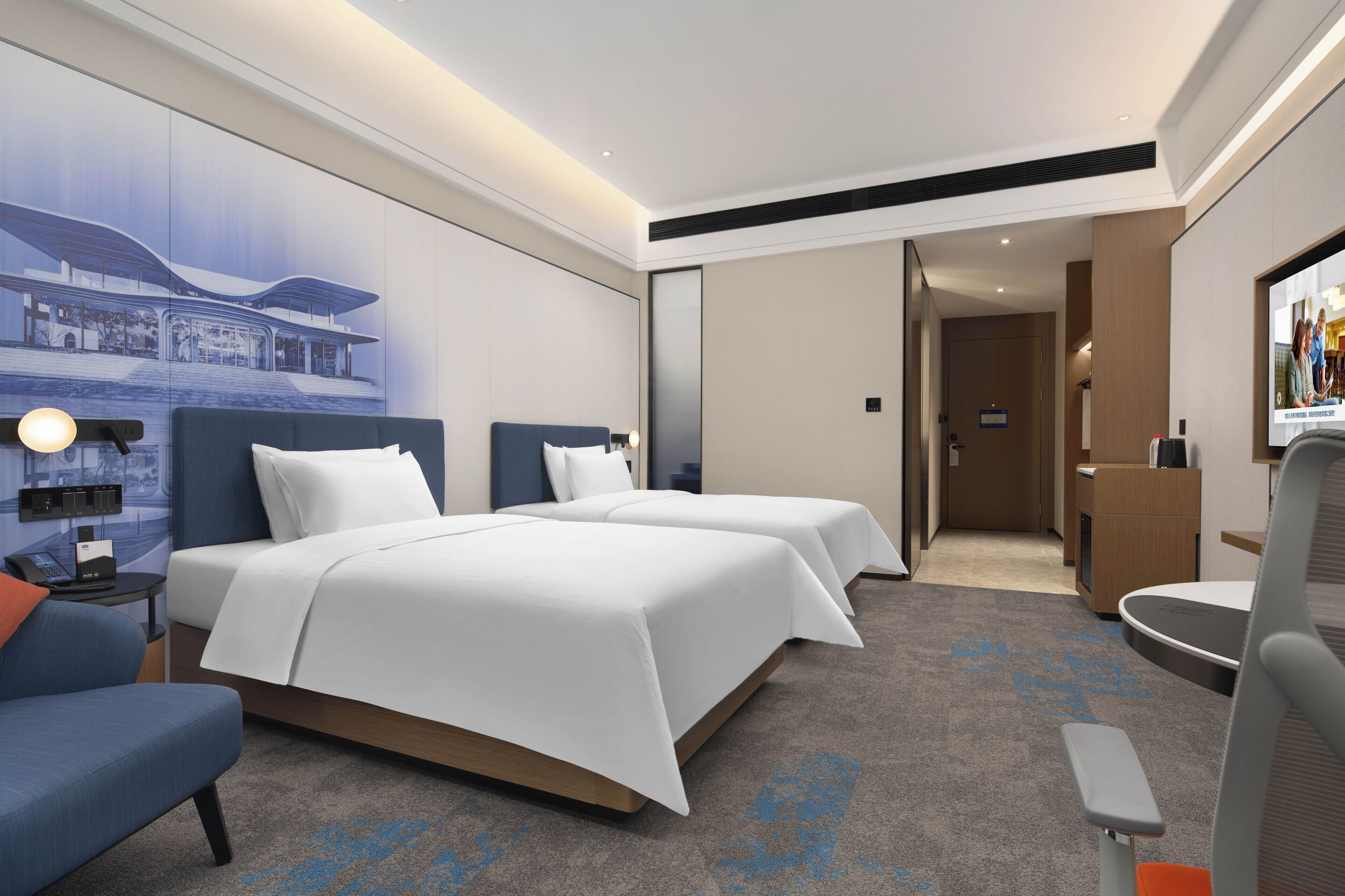 A hotel room with two beds, a desk, a chair, a desk lamp, and a wall-mounted television.