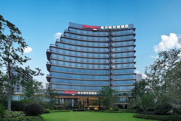 A modern hotel with a curved facade, multiple balconies, and a well-maintained garden.