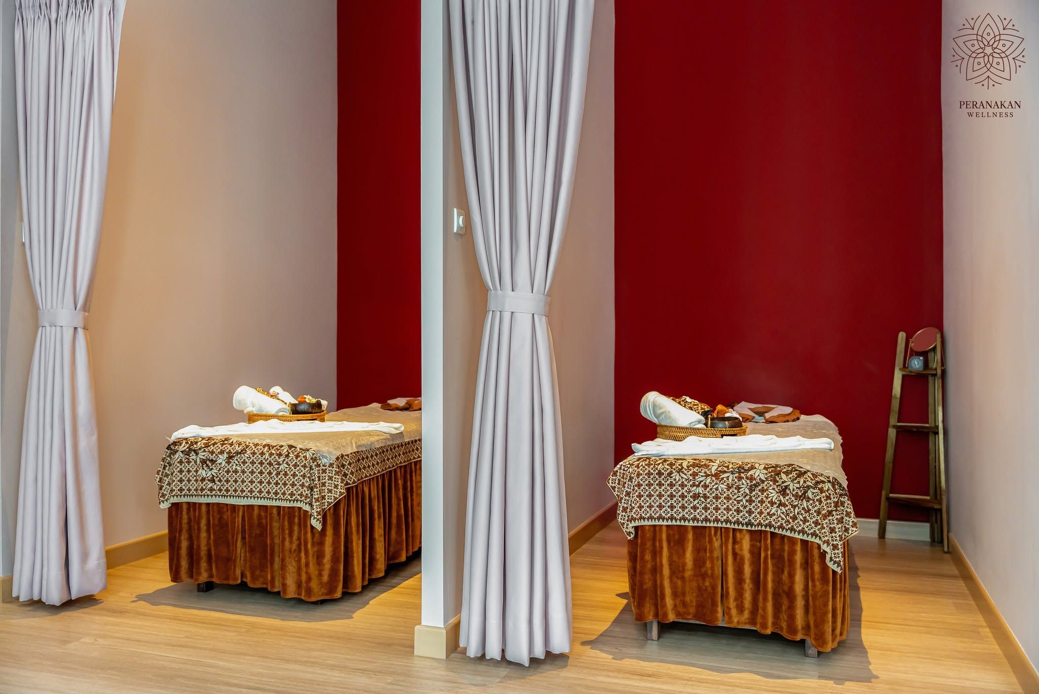 Twee massagetafels met handdoeken en een mand met spullen, voor een rode muur met witte gordijnen.