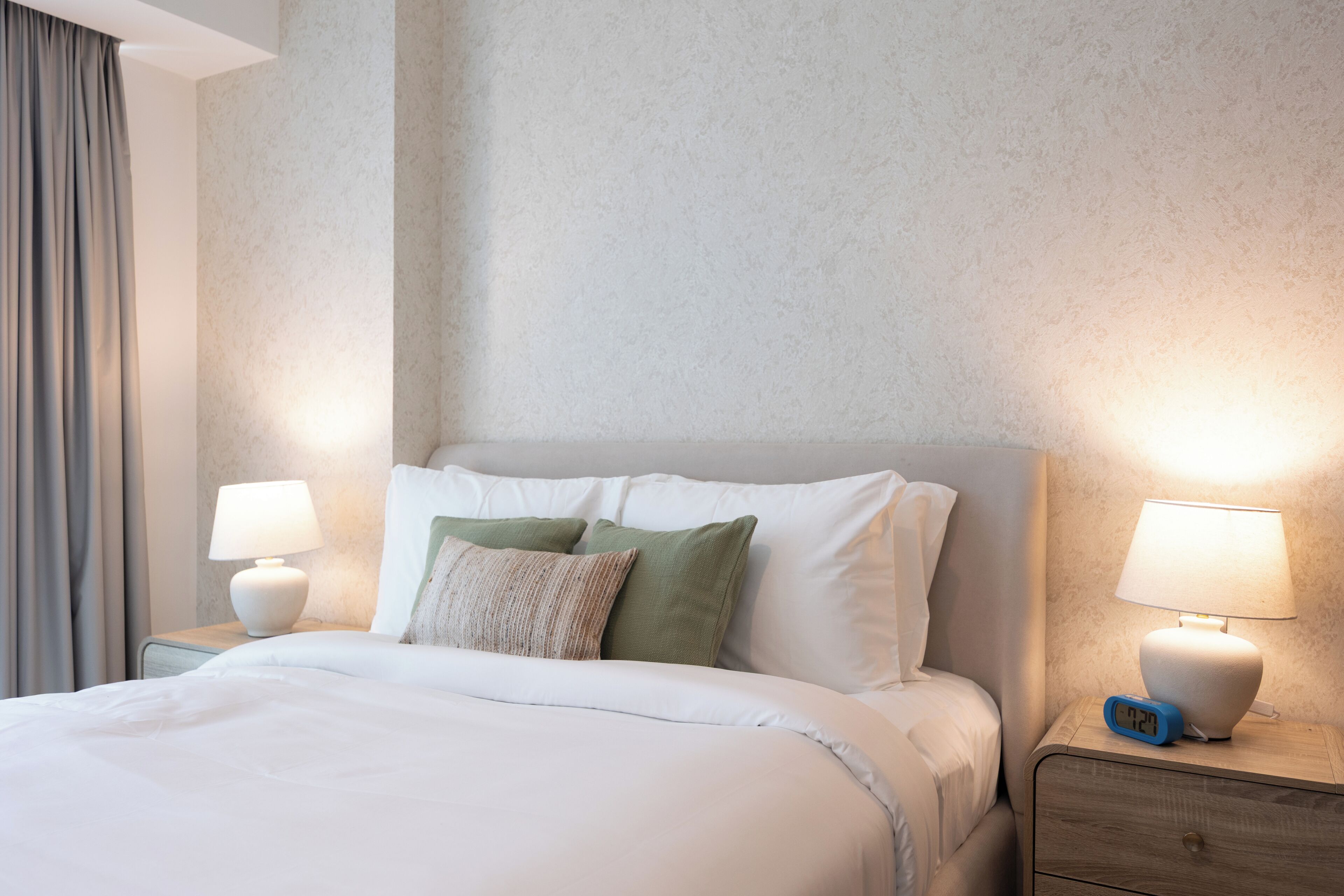 Une chambre d’hôtel comprenant un lit, deux tables de chevet, une petite table, une lampe et une horloge numérique.