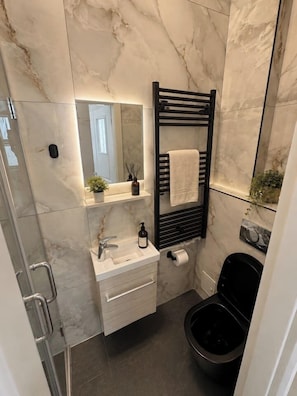 A bathroom with a marble wall, a black toilet, a white sink with a mirror above it, and a towel rack. - Contemporary Luxury Suite | Prime Central Location (Bucharest)