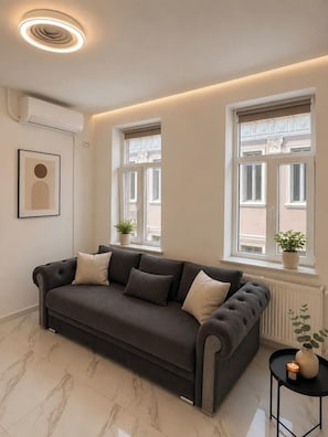 A modern living room with a dark grey sofa, two windows with white frames, a potted plant on each windowsill, and a small side table with a candle. - Contemporary Luxury Suite | Prime Central Location (Bucharest)