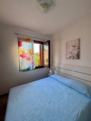 A bedroom with a bed, a window with curtains, a wall-mounted lamp, and a painting.