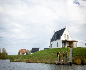 A modern house with a sloped roof, a dock extending into a body of water, and a grassy hill in the background. - Premium Water Villa in Thorn With Garden (Thorn)