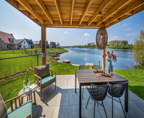 An outdoor patio with a wooden pergola, a table with chairs, a cushioned bench, and a view of a river and houses in the background. - Premium Water Villa in Thorn With Garden (Thorn)