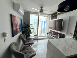 Un salon moderne doté d’un grand téléviseur, de sièges confortables, d’un ventilateur de plafond et offrant une vue sur de hauts immeubles par la fenêtre.