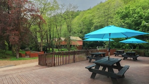A wooden deck with picnic tables and blue umbrellas, surrounded by trees and a wooden fence. - Cabin No. 4 - Salt and Fern Cabins (Port Talbot)