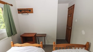 A small room with two wooden beds, a wooden table, a chair, a radiator, a window with a green curtain, and a wooden door. - Cabin No. 4 - Salt and Fern Cabins (Port Talbot)