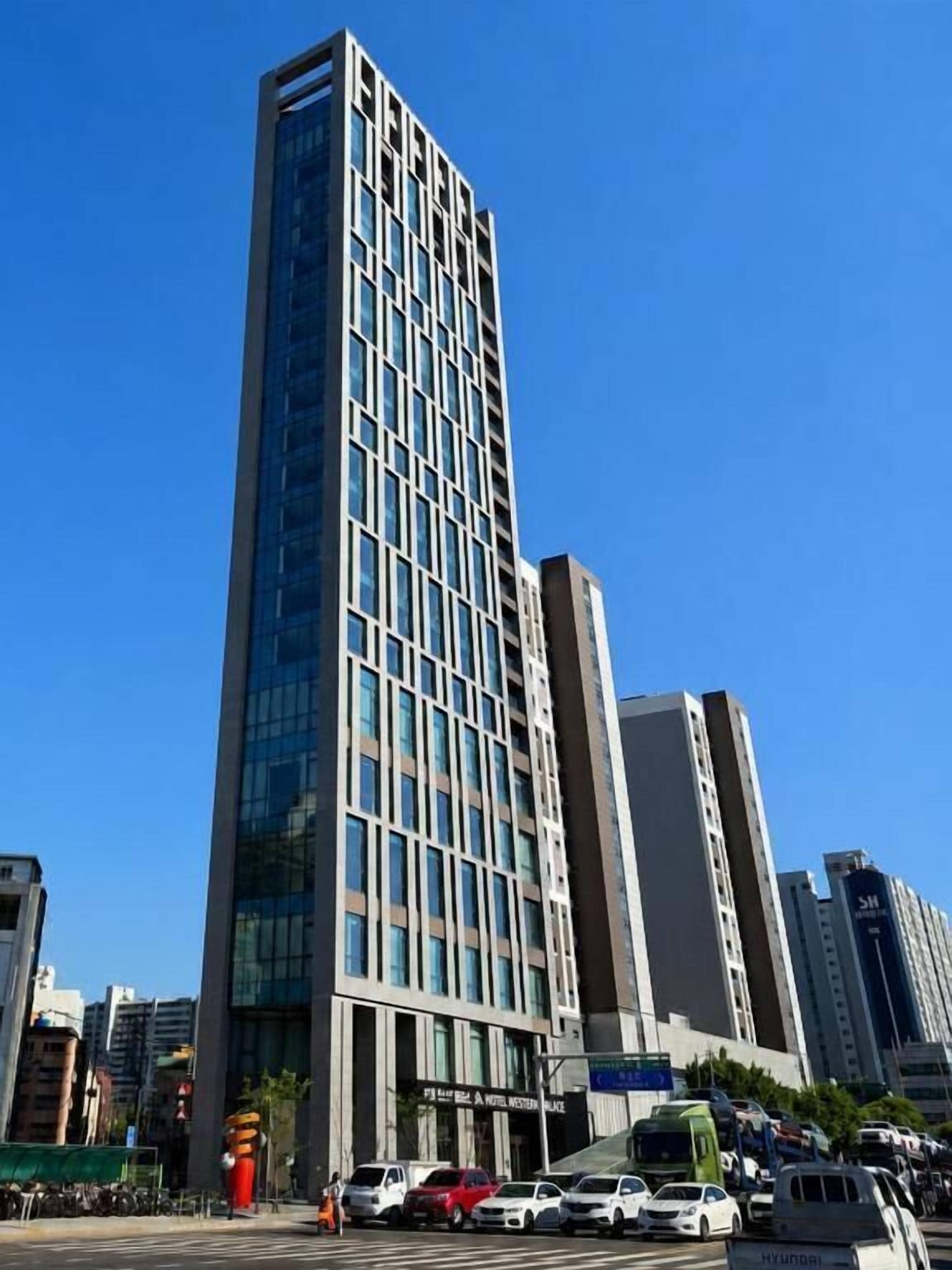 A tall, modern building with a grid of windows and a flat roof, surrounded by other high-rise buildings and a busy street with cars and a bus.