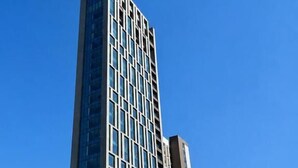 A tall, modern building with a grid of windows and a flat roof, surrounded by other high-rise buildings and a busy street with cars and a bus.