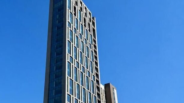 Un edificio alto y moderno con una fachada de ventanas enrejadas y un techo plano, rodeado de otros edificios de gran altura y una calle concurrida con coches y un autobús.