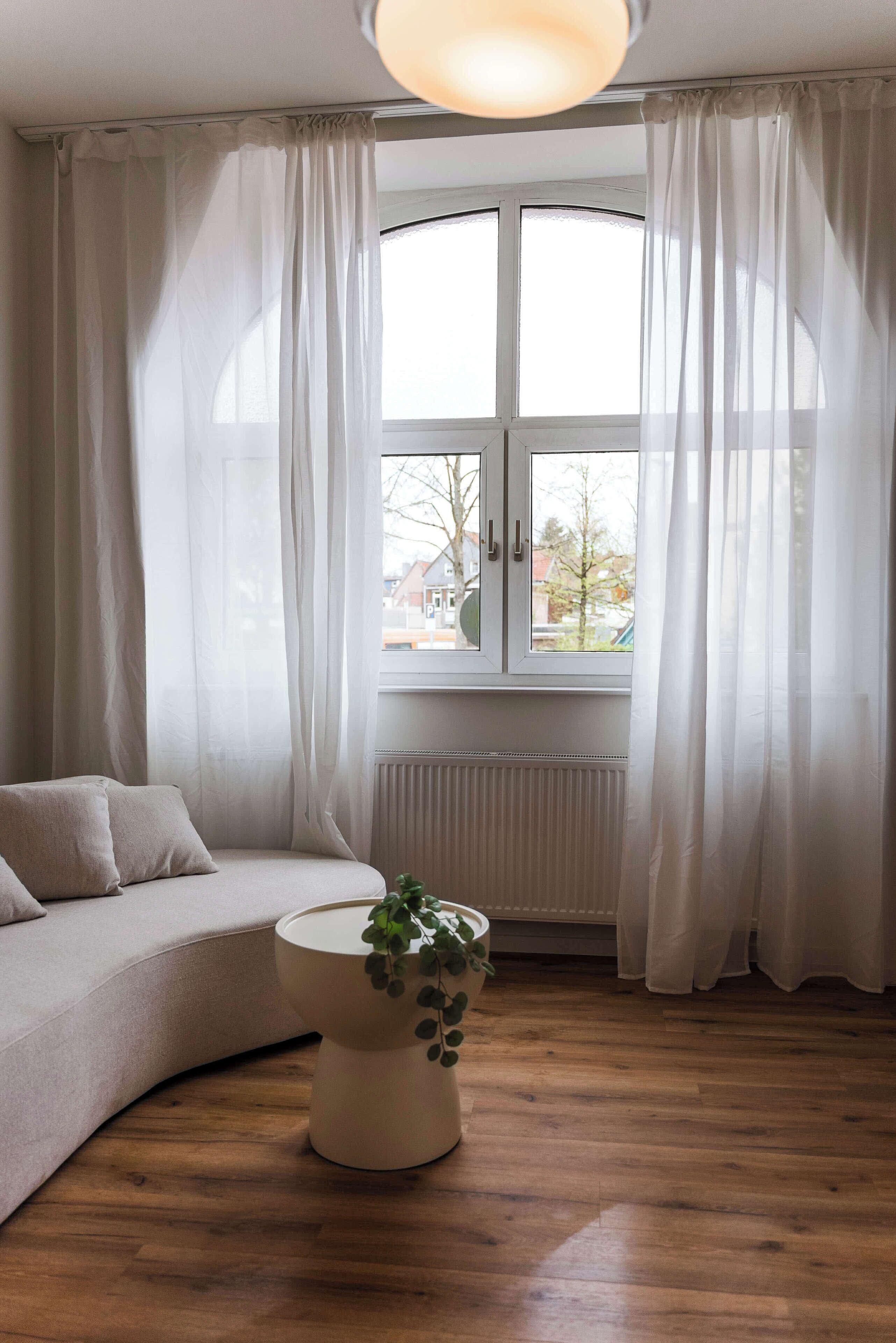 Ein Zimmer mit einer gebogenen Couch, einem runden Tisch mit einer Pflanze und einem Fenster mit weißen Vorhängen.