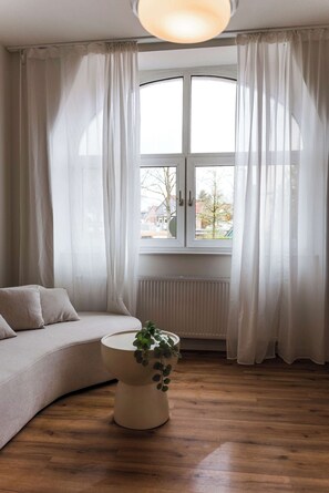 A room with a curved sofa, a round table with a plant, and a window with white curtains. - Design apartment by the park with parking (Celle)