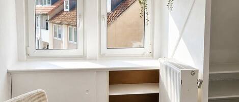 A room with a wooden dining table, two white upholstered chairs, a hanging plant, and a view of buildings through a window.