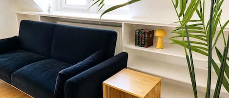 Una habitación con un sofá azul oscuro, una pequeña mesa de madera, una planta y un estante blanco con libros y una lámpara.