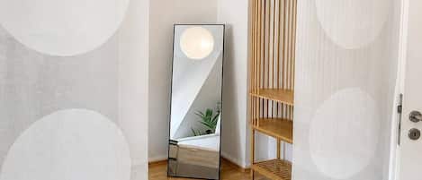 A hallway with a wooden floor, a white door, and a mirror reflecting a plant and a bench.