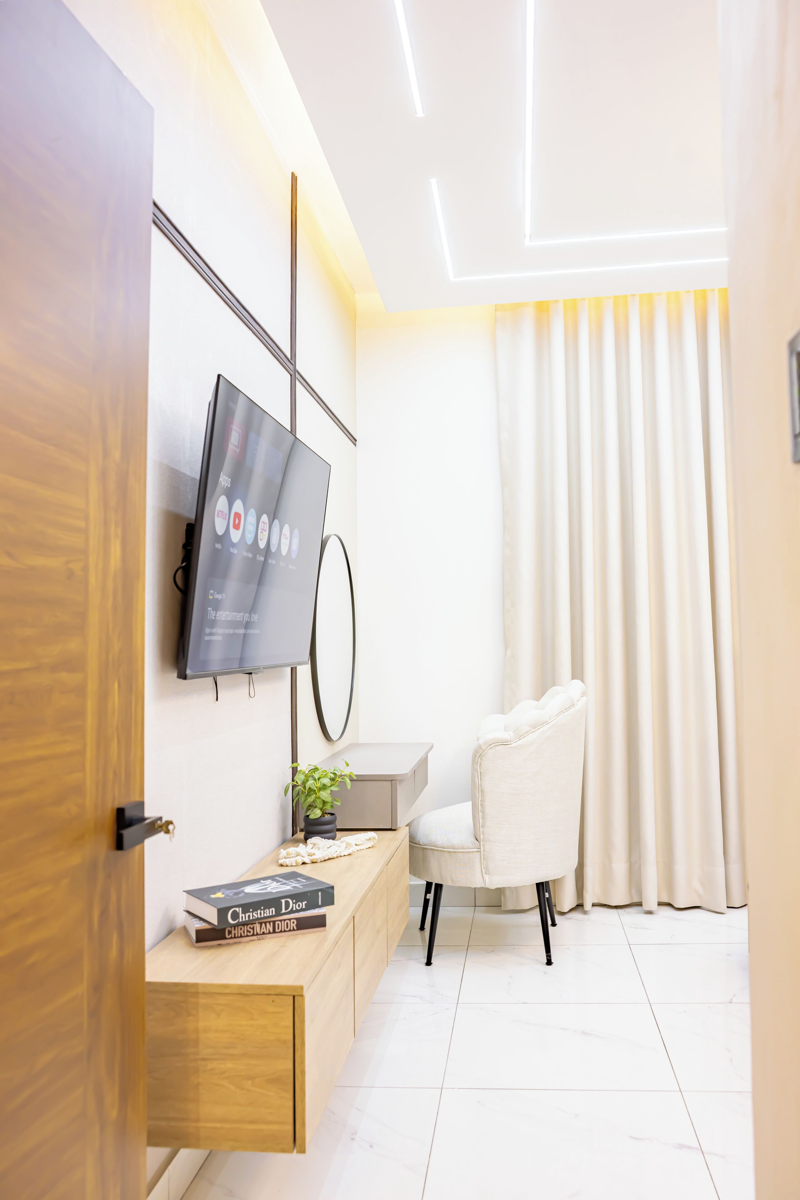 Ein modernes Hotelzimmer mit einem flachbildfernseher an der Wand, einem Holztischchen mit einem Buch, einer Topfpflanze und einem weißen Stuhl.
