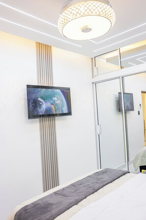 A modern bedroom with a flat-screen TV mounted on a wall, a bed with a grey headboard, and a large mirror reflecting the room. - Modern Luxury Apartment in Baní (Baní)