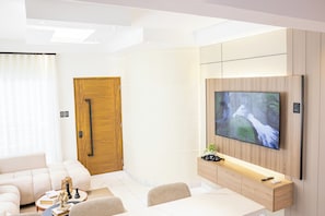 A modern living room with a flat-screen TV mounted on a wooden wall, a sofa, a coffee table with decorative items, and a door with a handle. - Modern Luxury Apartment in Baní (Baní)