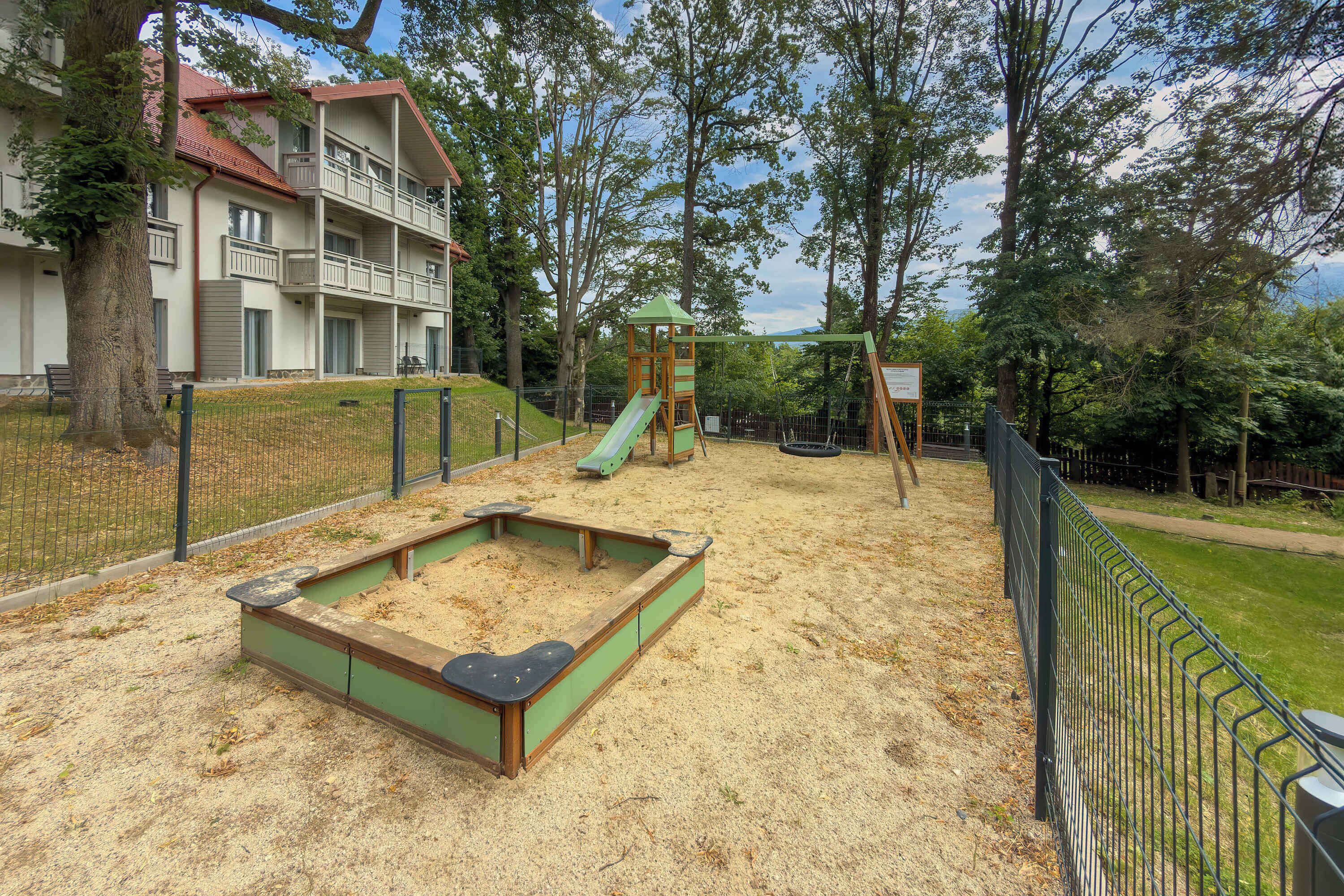 A playground with a slide and swings, surrounded by a fence and trees.
