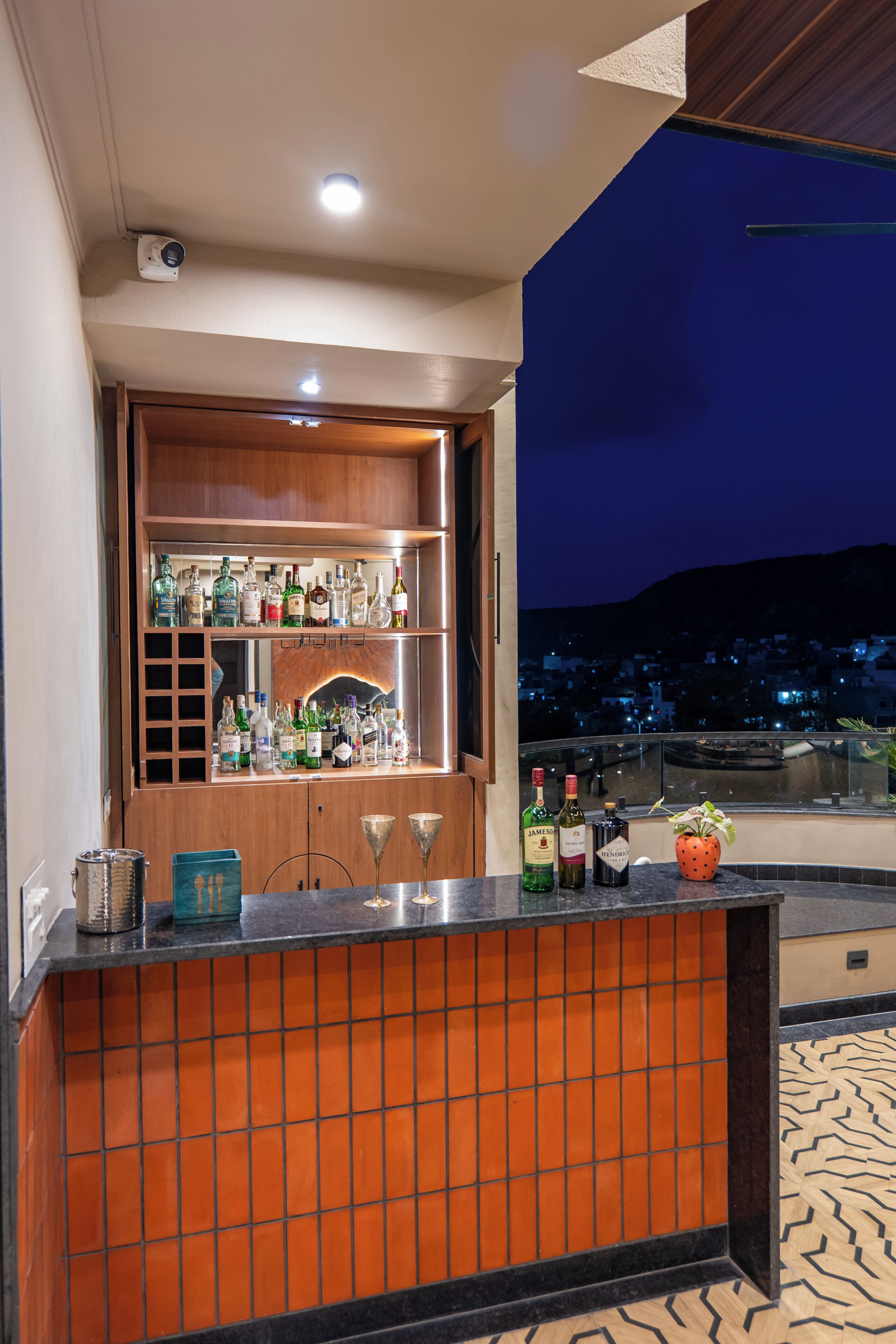 Eine Bar mit einem Holzboden voller Flaschen, einer Theke mit Gläsern und Blick auf eine beleuchtete Stadtlandschaft bei Nacht.