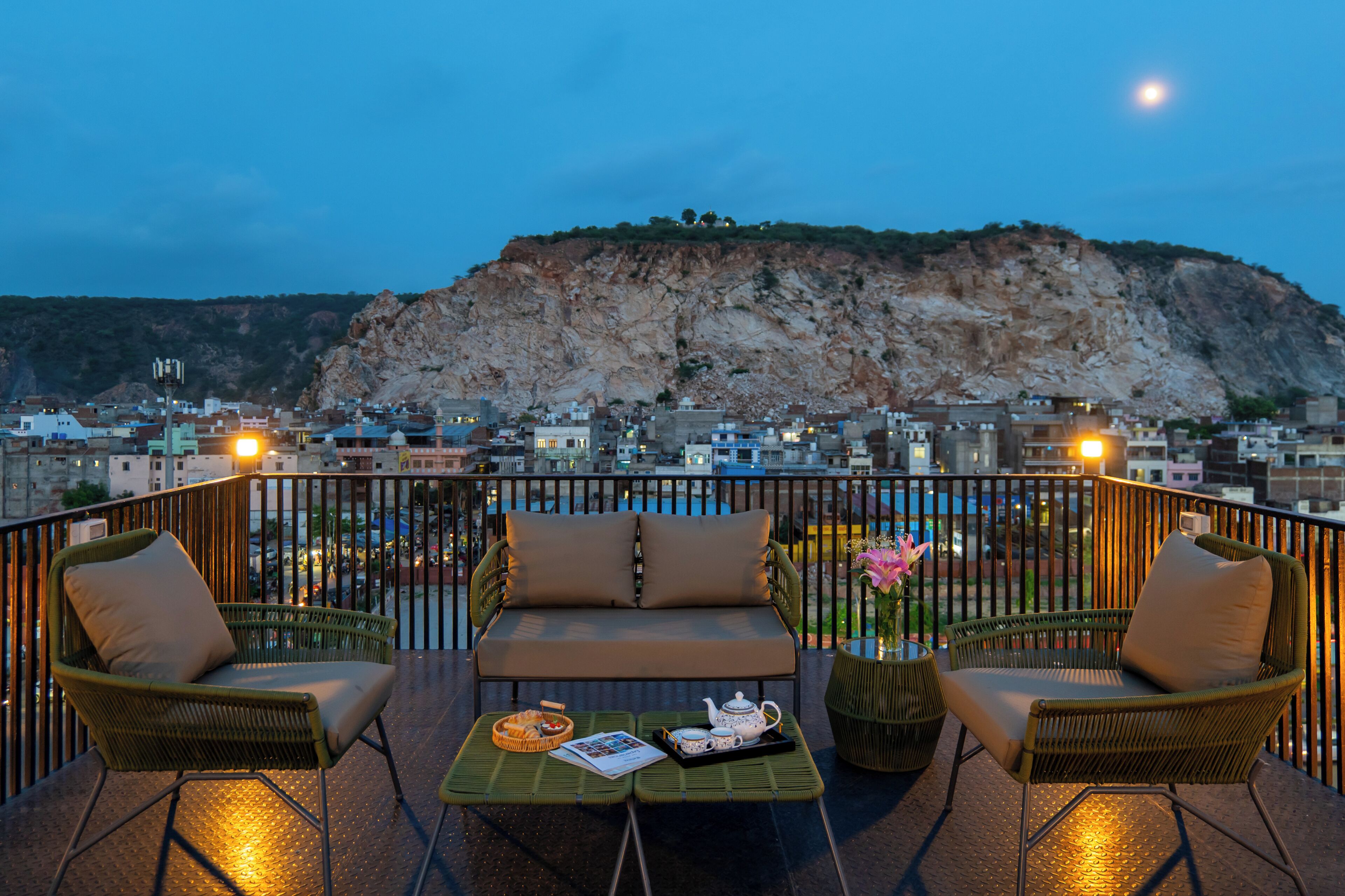 A balcony with wicker furniture, a table with a teapot and cups, and a view of a cityscape with a prominent cliff.