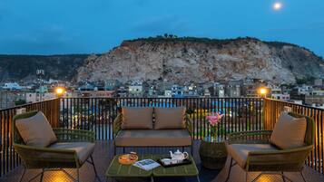 A balcony with wicker furniture, a table with a teapot and cups, and a view of a cityscape with a prominent cliff.