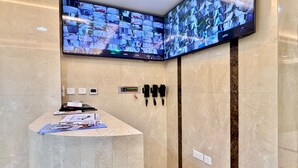 Una zona de recepción con mostrador de mármol, múltiples monitores mostrando imágenes de seguridad, un reloj digital y un sistema de intercomunicación instalado en la pared.