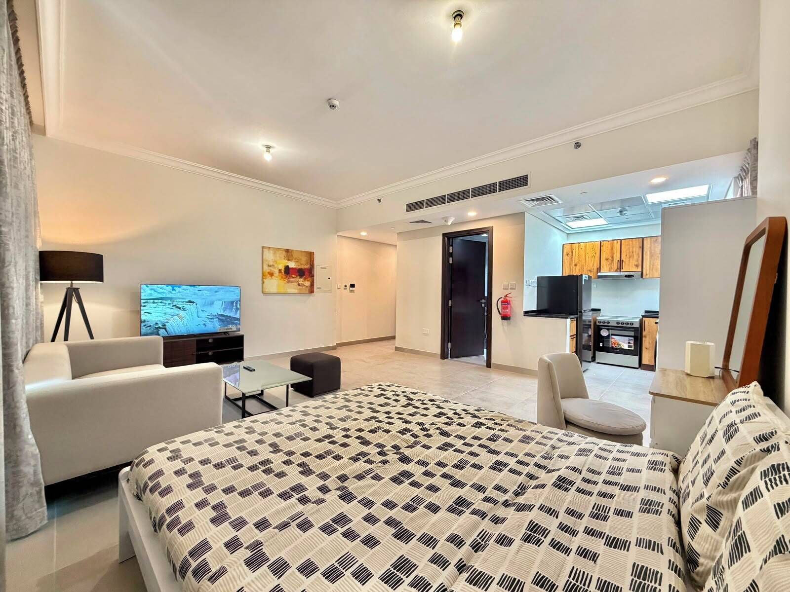 A modern hotel room with a large bed, a sofa, a TV, a desk, and a kitchenette.