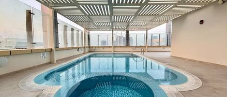 A rooftop swimming pool with a modern design, surrounded by glass walls and a metal ceiling structure.