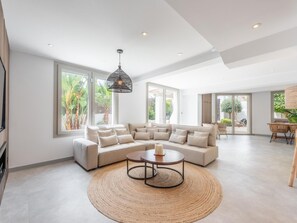 Ein geräumiges Wohnzimmer mit einer Eckcouch, einem runden Couchtisch und einem großen Fenster mit Blick ins Freie.